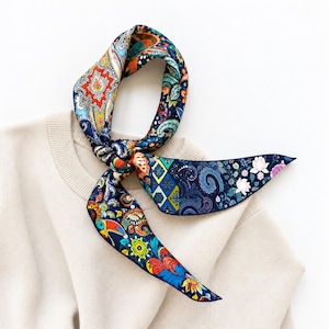 May include: A long, narrow silk scarf featuring a vibrant, multi-colored paisley and floral pattern on a dark blue background. The design includes intricate motifs, geometric shapes, and a toucan illustration, tied in a knot over a beige sweater.