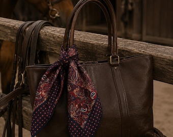 May include: A brown leather tote bag with a paisley scarf tied to the handle. The scarf has a navy blue border with white polka dots. The bag is sitting on a wooden fence, with a horse and tack in the background.