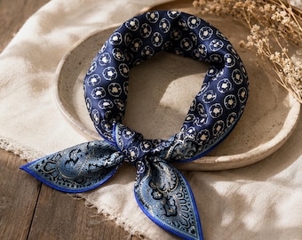 May include: A navy blue silk scarf with a white floral pattern and a paisley design on the ends. The scarf has a bright blue border and is tied in a knot. The scarf is displayed on a neutral-colored plate.