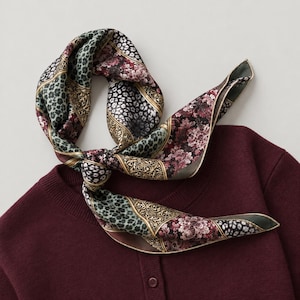 Op de afbeelding: Een lange, smalle sjaal met een patchworkontwerp van bloemen, luipaardprint en sierlijke gouden patronen in bordeauxrood, zwart en saliegroen, geknoopt en rustend op een effen bordeauxrood gebreid vest met knopen.