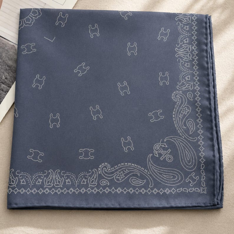 May include: A folded, square bandana in a muted blue hue. The fabric features a repeating pattern of small, white, stylized figures scattered across the surface. A detailed white paisley border frames the edges.