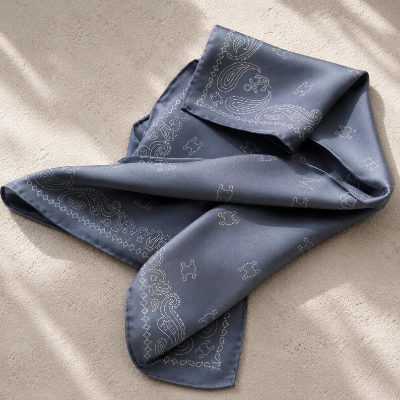 May include: A folded, slate blue silk scarf with a paisley and logo pattern. The scarf has a soft sheen and is folded to display the intricate design. The edges are finished with a delicate, white stitched border.