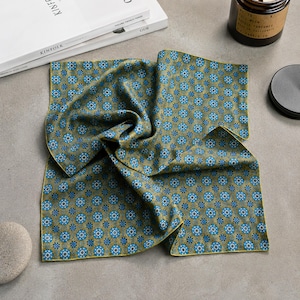 May include: A folded pocket square with a repeating floral pattern in shades of blue and gold. The edges are trimmed with a thin gold border. The fabric has a slight sheen, suggesting a silk or satin material. The pocket square is displayed on a neutral gray surface.