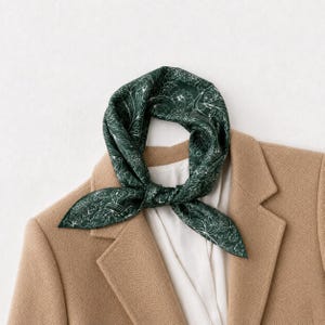 Op de afbeelding: Een donkergroene vierkante sjaal met een wit bloemmotief in lijntekening, gestrikt in een knoop. De sjaal ligt over een beige blazer met een wit overhemd eronder, wat zorgt voor een gladde textuur en een klassieke vouw.