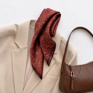 May include: A square scarf in a deep rust-red colour featuring a delicate white line-art floral pattern with a thin black border, draped over a beige blazer next to a smooth brown leather shoulder bag with a gold-toned metal zip and buckle.