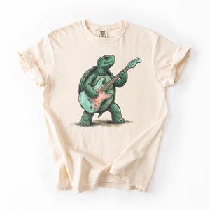 Puede incluir: Camiseta color crema con una tortuga verde tocando una guitarra eléctrica azul claro y rosa. La tortuga está de pie sobre sus patas traseras. La camiseta tiene cuello redondo clásico y mangas cortas.