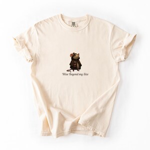 May include: A cream-coloured t-shirt with a cartoon mouse wearing a brown coat and hat. The text "Wise Beyond my Size" is printed below the mouse. The shirt has short sleeves and a crew neck.