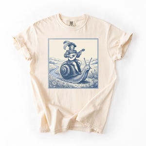 May include: A cream-coloured t-shirt with a blue and white illustration. The design shows a person playing a lute riding a snail. The image is framed within a blue border, set against a landscape with a winding path.