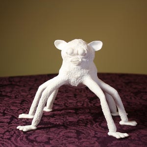 Könnte beinhalten: Eine weiße, strukturierte Skulptur einer Kreatur mit großen Augen, Ohren und mehreren beinartigen Anhängseln. Die Skulptur steht auf einer dunkelvioletten, gemusterten Oberfläche und einem gelbbraunen Hintergrund.