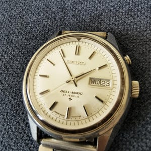 May include: A vintage Seiko Bell-Matic wristwatch with a silver dial and a stainless steel strap. The watch face shows the day and date, with "WED 23" visible. The watch has a silver case and a textured dial. The watch is a classic design.