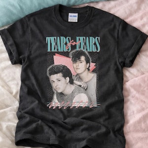May include: Black Gildan t-shirt featuring a retro design with the band name "Tears for Fears" in teal and pink. The shirt includes a black and white image of two men. The shirt is on a pink and blue background.