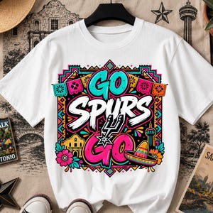 May include: A white short-sleeve t-shirt featuring a colorful graphic print with the text GO SPURS GO. The design includes a basketball team logo, a sombrero, a building silhouette, and floral patterns in bright pink, teal, and orange hues.