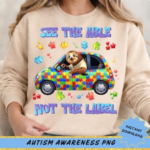 May include: Beige sweatshirt with a colorful graphic featuring a sloth driving a car made of puzzle pieces. Text reads "SEE THE ABLE NOT THE LABEL" in blue. Additional puzzle pieces and stars surround the car. "AUTISM AWARENESS PNG" is at the bottom.