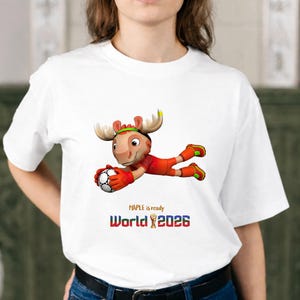 Puede incluir: Camiseta blanca con un personaje de dibujos animados de un alce con uniforme de f&uacute;tbol, atrapando un bal&oacute;n de f&uacute;tbol. El texto "MAPLE is ready World 2026" est&aacute; impreso debajo en azul, rojo y dorado.
