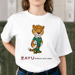 May include: White t-shirt featuring a cartoon jaguar mascot holding a soccer ball. The jaguar wears a green and white soccer uniform. The text "ZAYU WORLD CUP 2026" is printed in red and black below the jaguar.