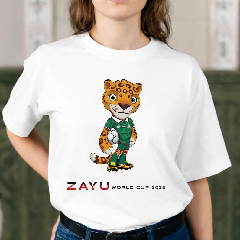 May include: White t-shirt featuring a cartoon jaguar mascot holding a soccer ball. The jaguar wears a green and white soccer uniform. The text "ZAYU WORLD CUP 2026" is printed in red and black below the jaguar.