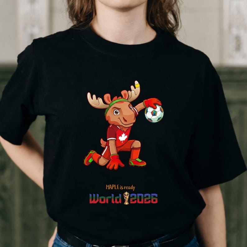 Puede incluir: Camiseta negra con un alce de dibujos animados con uniforme de f&uacute;tbol, sosteniendo un bal&oacute;n de f&uacute;tbol. El alce lleva una camiseta roja con una hoja de arce. El texto en la camiseta dice "MAPLE is ready World 2026".