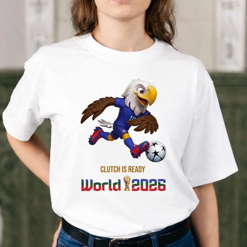 Puede incluir: Camiseta blanca con un &aacute;guila de dibujos animados con uniforme de f&uacute;tbol azul, pateando un bal&oacute;n. El texto "CLUTCH IS READY" y "World 2026" est&aacute; impreso debajo.