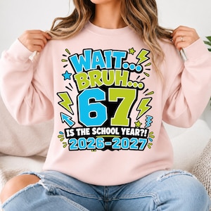 May include: A light pink crewneck sweatshirt featuring a graphic print with the text "WAIT... BRUH... 67 IS THE SCHOOL YEAR?! 2026-2027". The design includes bright blue and lime green lettering, numbers, and lightning bolt accents on the front.