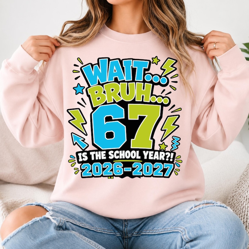 May include: A light pink crewneck sweatshirt featuring a graphic print with the text "WAIT... BRUH... 67 IS THE SCHOOL YEAR?! 2026-2027". The design includes bright blue and lime green lettering, numbers, and lightning bolt accents on the front.