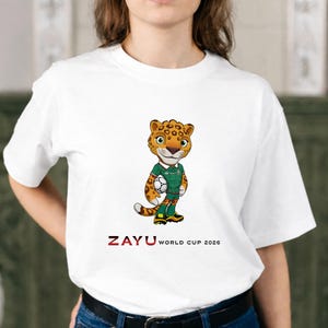 Puede incluir: Camiseta blanca con una mascota de jaguar de dibujos animados sosteniendo un bal&oacute;n de f&uacute;tbol. El jaguar lleva un uniforme verde y blanco. El texto "ZAYU WORLD CUP 2026" est&aacute; impreso en rojo debajo.