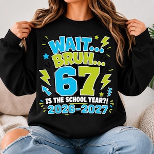 May include: A black long-sleeve sweatshirt featuring a graphic print with the text "WAIT... BRUH... 67 IS THE SCHOOL YEAR?! 2026-2027". The design includes large blue and lime green numbers and letters, accented by small lightning bolt and star shapes.