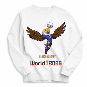 Puede incluir: Sudadera blanca con una mascota de &aacute;guila de dibujos animados con uniforme de f&uacute;tbol azul, equilibrando un bal&oacute;n de f&uacute;tbol. El texto "CLUTCH IS READY" y "World 2026" est&aacute;n impresos debajo, con un trofeo de la Copa del Mundo.