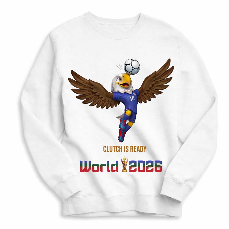 Puede incluir: Sudadera blanca con una mascota de &aacute;guila de dibujos animados con uniforme de f&uacute;tbol azul, equilibrando un bal&oacute;n de f&uacute;tbol. El texto "CLUTCH IS READY" y "World 2026" est&aacute;n impresos debajo, con un trofeo de la Copa del Mundo.