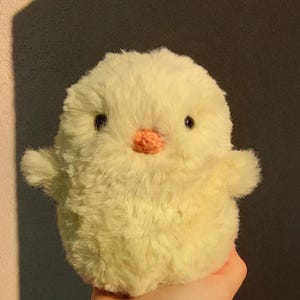 May include: A round, fluffy, pale yellow plush toy shaped like a small chick. The toy features a soft, textured faux fur exterior, two small dark circular eyes, and a tiny, rounded, light orange crocheted beak centred on the face.