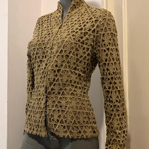 Puede incluir: Chaqueta calada de color beige con cuello alto y mangas largas. La chaqueta presenta un estampado floral repetido en toda la tela. La prenda se exhibe en un maniquí.