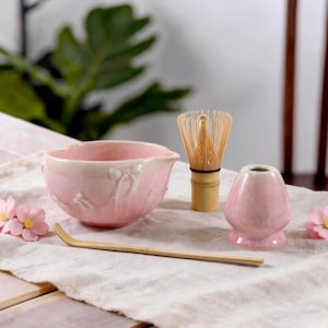 Op de afbeelding: Een lichtroze keramische matchakom met reliëf bloemdetails, een bamboe garde, een bamboe lepel en een bijpassende roze keramische gardehouder. De set is gerangschikt op een lichtgekleurde stoffen doek, vergezeld door twee kleine roze kunstbloemen.