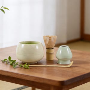 May include: A matcha tea set on a wooden table. The set includes a light green ceramic bowl, a bamboo whisk, a small light green ceramic stand, and a bamboo scoop. The bowl contains green tea. A sprig of greenery is on the table.