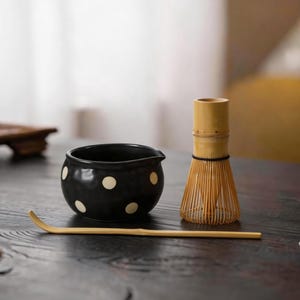 Può includere: Una ciotola in ceramica nera con pois bianchi, un frustino per matcha in bambù di colore chiaro con un filo nero avvolto e un cucchiaio da tè in bambù lungo e sottile, disposti insieme su una superficie in legno scuro per preparare il tè giapponese.