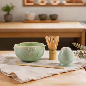 May include: A light green ceramic matcha bowl with embossed bow details, a bamboo whisk, a bamboo tea scoop, and a matching green ceramic whisk holder. The set is arranged on a light-coloured fabric cloth, showcasing a soft, glazed finish.