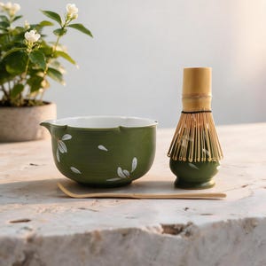 May include: A green ceramic matcha bowl with white floral accents, a bamboo whisk resting on a matching green ceramic stand, and a long, thin bamboo tea scoop. The bowl features a small pouring spout and a smooth, white interior finish.