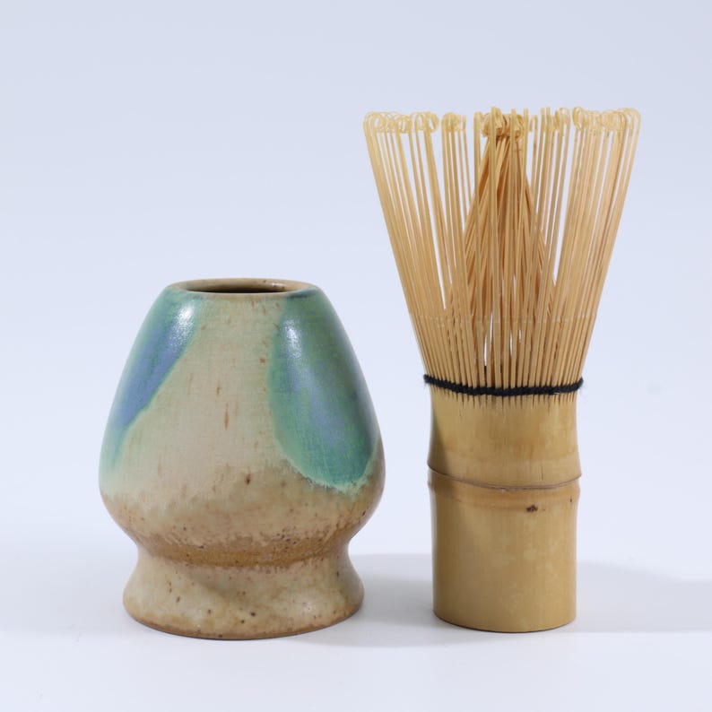 May include: A ceramic matcha bowl with blue and green glaze and a bamboo matcha whisk. The bowl is a light beige color with a rounded shape. The whisk is made of thin bamboo tines bound together with a black thread.
