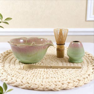 Puede incluir: Un juego de té matcha con un cuenco de cerámica verde y rosa, un batidor de bambú, un pequeño soporte de cerámica y una cuchara de bambú. El juego está dispuesto sobre un salvamanteles tejido de color crema.