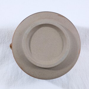 May include: A ceramic plate with a speckled, tan surface. The plate has a raised, circular center and a wide, flat rim. The rim and the edge of the plate are a darker brown color. The plate is likely handmade.