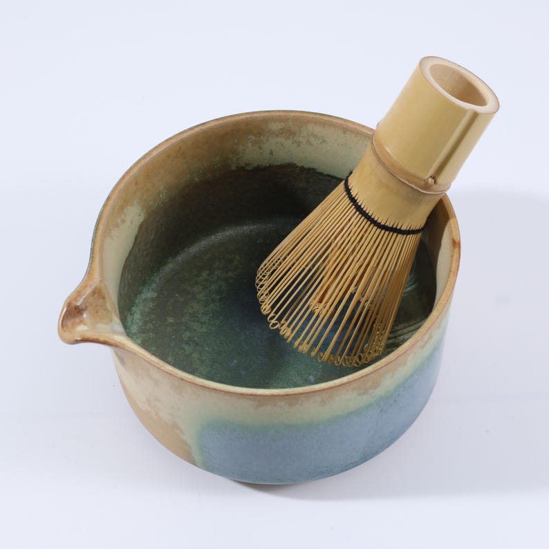 May include: A ceramic matcha bowl with a spout, featuring a gradient of colors from beige to blue-green. A bamboo matcha whisk rests inside the bowl, ready for use. This set is ideal for preparing traditional matcha tea.