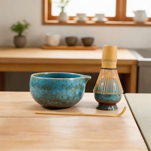 Może przedstawiać: Turkusowa ceramiczna miska do matchy z dzióbkiem, bambusowa trzepaczka i bambusowa łyżka spoczywają na jasnej drewnianej powierzchni. Trzepaczka ma bambusowy uchwyt i włosie. Miska i trzepaczka mają niebieskie i brązowe szkliwo.