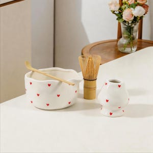 May include: A white ceramic matcha bowl with a scalloped rim and small red heart patterns, accompanied by a light brown bamboo whisk, a bamboo scoop, and a matching white ceramic whisk holder decorated with red hearts.