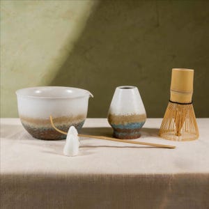 May include: A matcha tea set featuring a white and brown ceramic bowl with a spout, a white ceramic water scoop, a bamboo whisk, and a ceramic tea container. The set is arranged on a beige linen tablecloth.