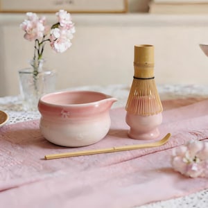 May include: A pink and white ceramic tea bowl, a bamboo whisk, and a bamboo scoop sit on a pink cloth. The tea bowl has a floral design. A small vase of pink flowers is in the background.