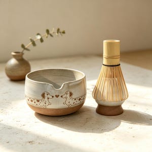 Puede incluir: Un cuenco de cerámica para matcha con pico vertedor, decorado con ilustraciones de perros salchicha con manchas marrones y un pequeño corazón. Al lado hay un batidor de bambú y un pequeño jarrón de cerámica beige con una rama verde.
