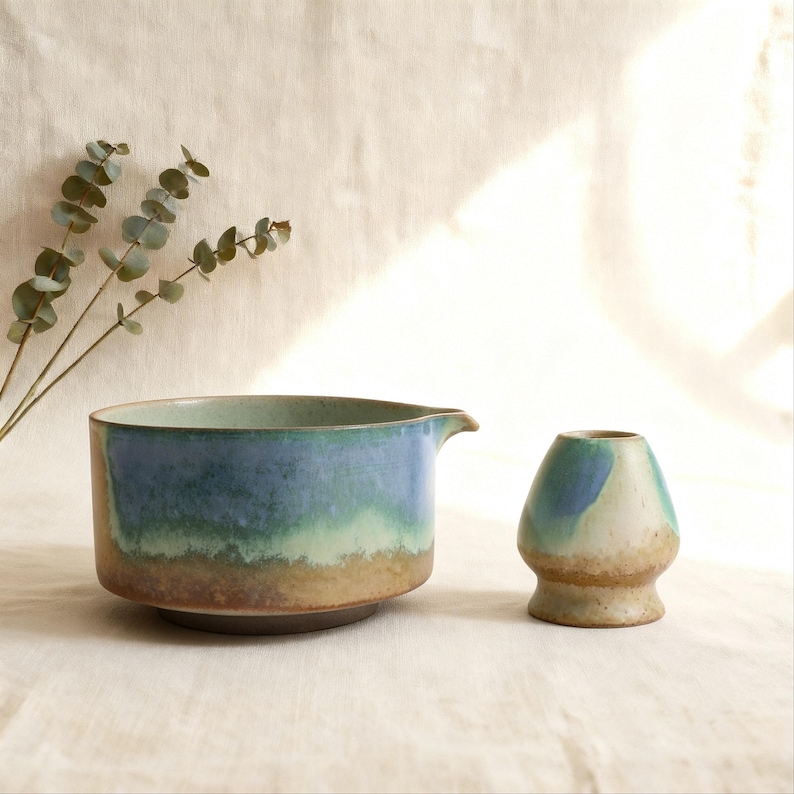 May include: Ceramic tea set featuring a blue and green gradient glaze. The set includes a wide-mouthed bowl with a spout and a small, rounded vessel. Eucalyptus sprigs add a natural touch to the composition.