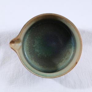 May include: A handmade ceramic bowl with a dark green interior and a light brown rim. The bowl has a small handle on one side. The interior has a speckled texture, and the rim is a lighter shade of brown. The bowl is sitting on a white surface.