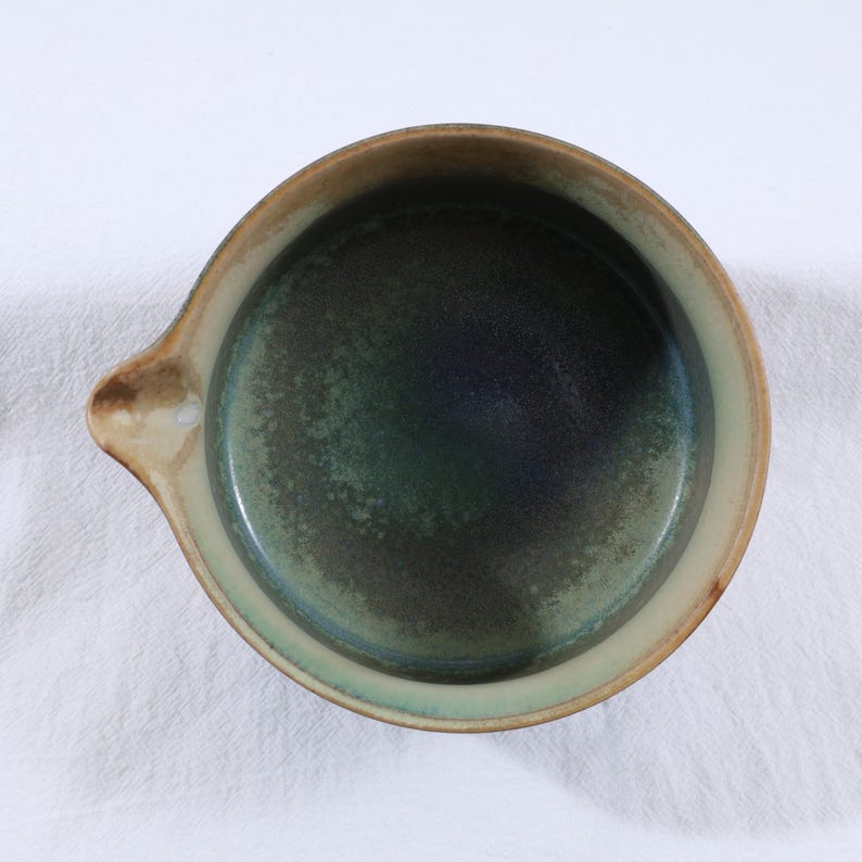 May include: A handmade ceramic bowl with a dark green interior and a light brown rim. The bowl has a small handle on one side. The interior has a speckled texture, and the rim is a lighter shade of brown. The bowl is sitting on a white surface.