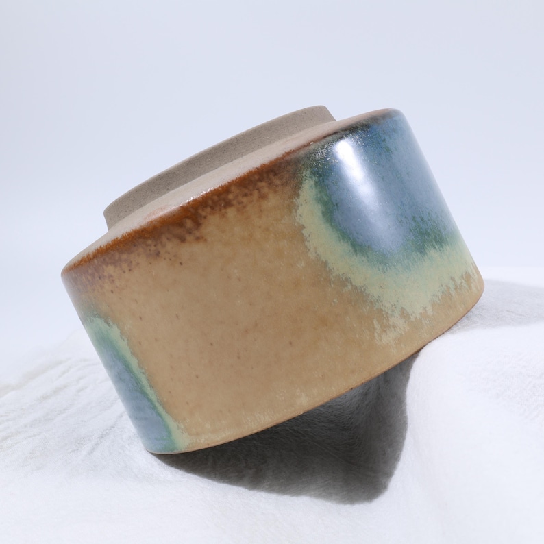May include: A ceramic jar with a lid. The jar has a cylindrical shape with a wide base and a narrower top. The exterior features a blend of colors, including tan, blue, and green, creating a unique, artistic appearance. The lid is a solid tan color.