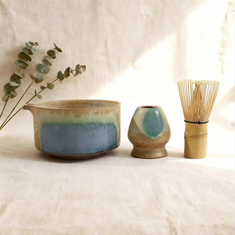 May include: A ceramic matcha bowl with blue and tan glaze, a small ceramic tea caddy, and a bamboo whisk. The bowl has a spout. A sprig of eucalyptus is in the background. The items are on a neutral-colored surface.