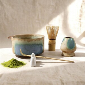 May include: A matcha tea set featuring a ceramic bowl with blue and brown glaze, a bamboo whisk, a whisk holder, a scoop, and a small ceramic container. A pile of green matcha powder is also present.